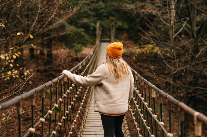 Eine Person mit gelber Mütze und beigem Mantel überquert eine Hängebrücke in einem herbstlichen Wald. Sie steht mittig auf der Brücke, umgeben von kahlen, teils gelb belaubten Bäumen.