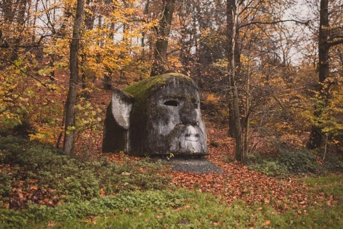 Große, moosbedeckte Steinfigur eines Gesichtes im Wald. Umgeben von buntem Herbstlaub und Bäumen.