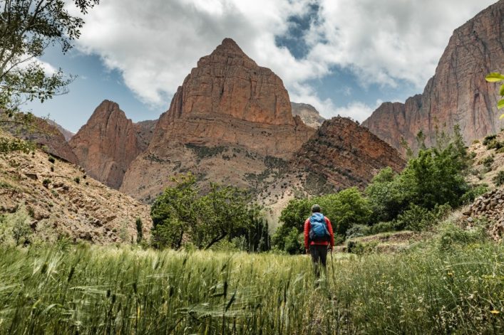 In einem grünen Tal steht ein Wanderer mit blauem Rucksack und roter Jacke, umgeben von Gras und Bäumen. Im Hintergrund ragen majestätische, felsige Berge unter bewölktem Himmel auf.