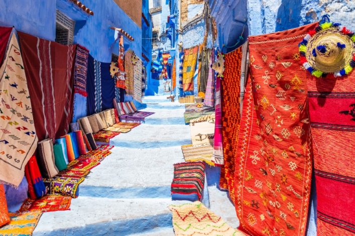 Enge Gasse mit blauen Wänden in Chefchaouen, gesäumt von bunten Teppichen und Kissen auf beiden Seiten. Die Souvenirs erzeugen ein lebhaftes und farbenfrohes Ambiente.