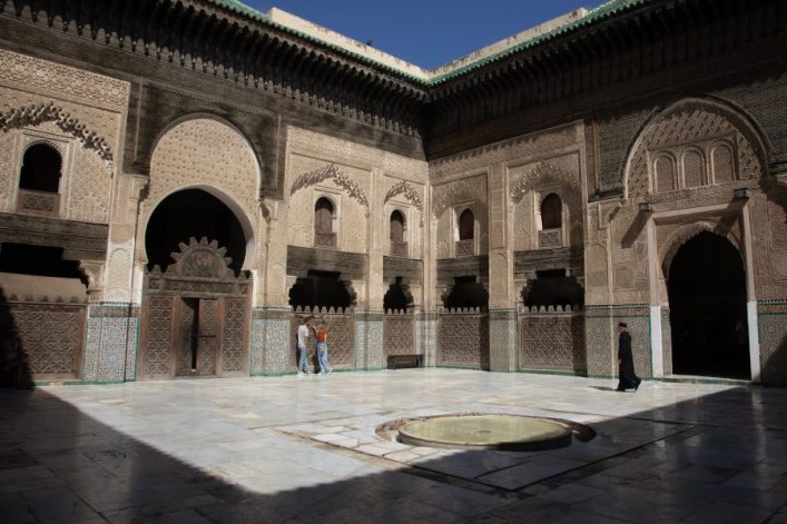 Ein sonniger Innenhof mit kunstvoll verzierten Wänden und Bögen in beige und grün. Zwei Personen gehen links, eine rechts. In der Mitte des Marmorbodens befindet sich ein flaches, rundes Becken.