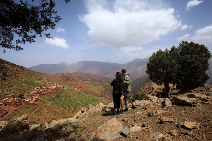 Auf dem Bild stehen zwei Personen auf einem felsigen Aussichtspunkt. Links erstrecken sich terrassierte Felder und ein Dorf, rechts ragt ein einzelner Baum empor. Im Hintergrund sind Berge und ein bewölkter Himmel.