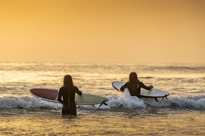 Zwei Menschen in Neoprenanzügen tragen Surfbretter ins Meer. Die untergehende Sonne taucht die Szene in warmes Licht, der Ozean erstreckt sich bis zum Horizont.