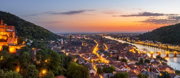 Heidelberger Schloss erstrahlt links im Abendlicht. In der Mitte schlängelt sich ein Fluss durch die Stadt. Rechts erhebt sich ein bewaldeter Hügel, während der Himmel in warmen Farben leuchtet.