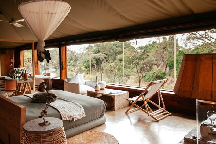 Gemütliche Safari-Lodge mit großem Bett, Holzstühlen und Panoramafenster. Auf der rechten Seite Sessel und Tisch, draußen grüne Landschaft. Heller Raum mit Holzelementen und stilvoller Dekoration.