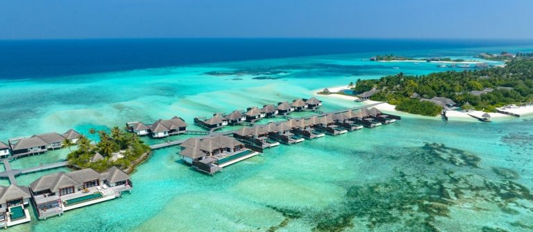 Über türkisfarbenem Wasser schweben Wasserbungalows auf Holzstegen, umgeben von dicht bewaldeten Inseln und einem strahlend blauen Himmel im Hintergrund.