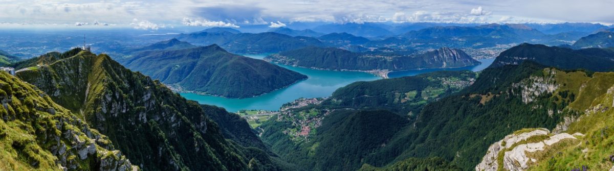 Blick von Monte Generoso im Tessin: Vorne schroffe Felsen und grüne Hügel, dazwischen ein türkisfarbener See. Im Hintergrund ziehen sich Berge bis zum Horizont unter einem bewölkten Himmel.