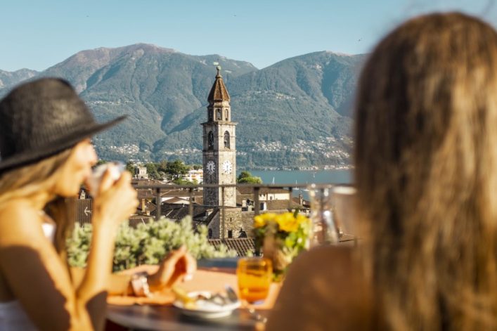 Eine Frau mit Hut trinkt Kaffee, im Hintergrund ein Kirchturm und Berge. Ein Tisch mit Getränken und Blumen ist im Vordergrund, der See schimmert in der Mitte. Links der Turm, rechts die Frau von hinten.