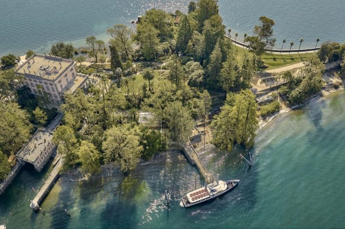 Blick von oben auf die üppig bewaldeten Brissago-Inseln im blauen Wasser eines Sees. Ein Boot ankert an einem Steg im Vordergrund. Rechts schlängelt sich ein Weg mit Palmen am Ufer entlang.