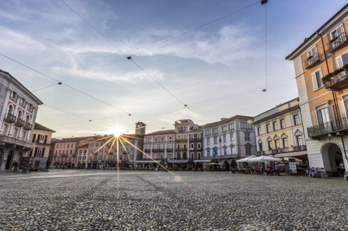 Steinpflasterplatz in Locarno bei Sonnenuntergang; umgeben von bunten historischen Gebäuden, Cafétische und Stühle rechts im Vordergrund, leere Piazza zieht sich bis zum Horizont, vorbei an Arkaden.