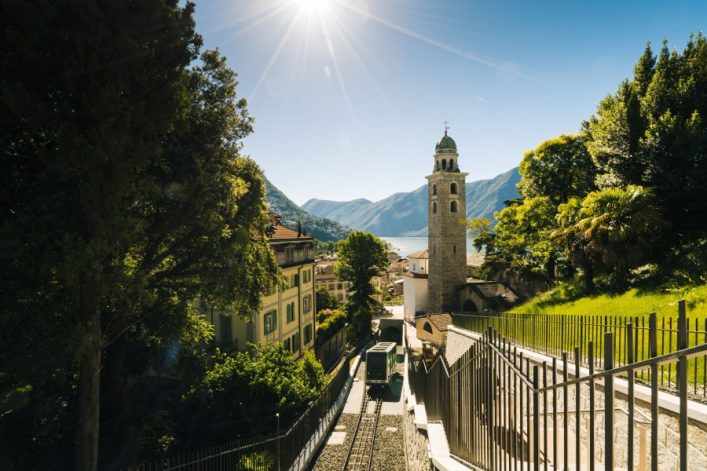 Blick auf Lugano mit sonnigem Himmel. Im Vordergrund Schienen zwischen Bäumen, links Gebäude. Zentral ein Turm, im Hintergrund Berge und Wasser. Klare, helle Atmosphäre.