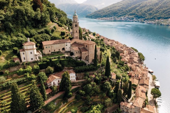 Morcote, Schweiz: Historisches Dorf am Seeufer mit alten Gebäuden, einer Kirche mit Turm zentral. Üppige Vegetation und Terrassen mit Zypressen führen zur malerischen Küstenlinie, umgeben von Bergen.