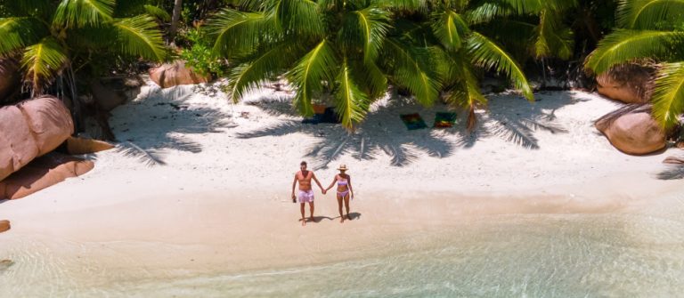 Zwei Personen in Badekleidung stehen Hand in Hand auf einem weißen Sandstrand. Hinter ihnen Palmen, davor das klare blaue Meer. Große Felsen umgeben den idyllischen Strandabschnitt.
