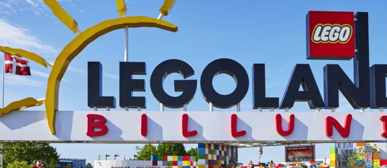 Das Bild zeigt den bunten LEGOLAND Billund Eingang mit großen LEGO-Buchstaben, Zahnrädern, einer Sonne und einer dänischen Flagge im Hintergrund.