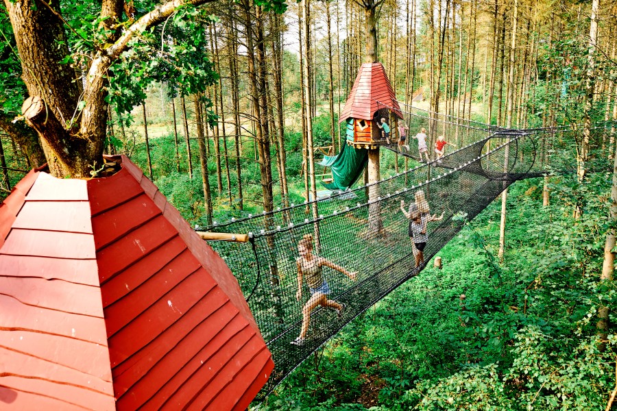 Netzbrücke verbindet zwei Baumhäuser mit roten Dächern in einem Wald. Kinder klettern darauf. Üppiges grünes Laub umgibt die Szene. Eines der Baumhäuser ist vorne links, das andere zentral.