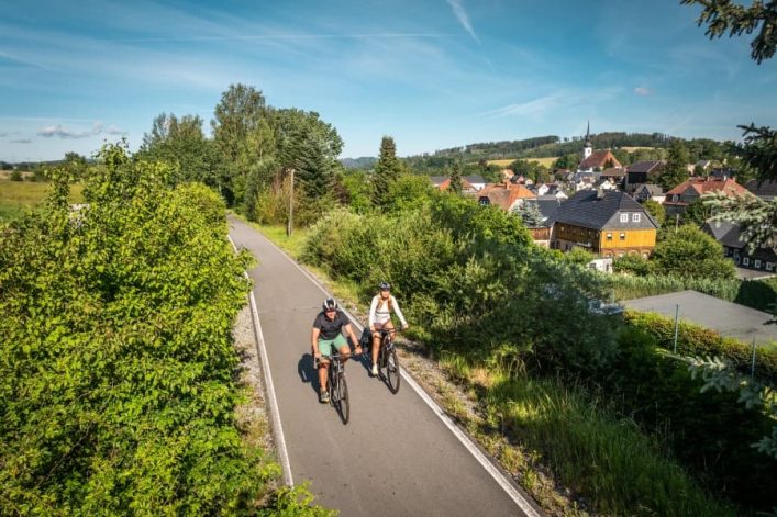 Zwei Radfahrer auf einem asphaltierten Weg, umgeben von grüner Vegetation. Im Hintergrund ein Dorf mit traditionellen Häusern und einem Kirchturm unter blauem Himmel. Links Bäume, rechts Büsche.
