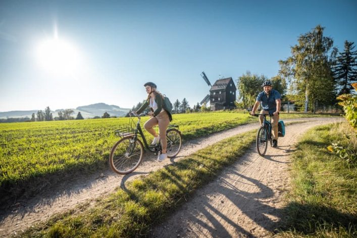 Zwei Radfahrer radeln auf einem Feldweg in ländlicher Umgebung. Links erscheint die Sonne am Himmel. Im Hintergrund steht eine Holz-Windmühle, rechts säumen Bäume den Weg.
