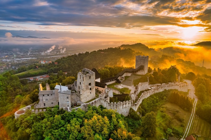 Eine große Burganlage mit Mauern und Türmen liegt auf einem bewaldeten Hügel, während die Sonne über einer nebligen Landschaft mit Stadt und Feldern aufgeht.