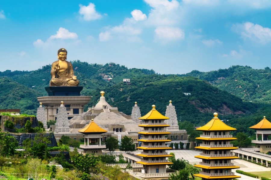 Das Bild zeigt den Fo-Guang-Shan-Buddha-Komplex in Taiwan. Im Vordergrund befinden sich mehrere Pagoden mit goldenen Dächern, während im Hintergrund eine riesige goldene Buddha-Statue über der Tempelanlage thront. Die Szenerie ist von grünen Hügeln und einem klaren blauen Himmel umgeben.