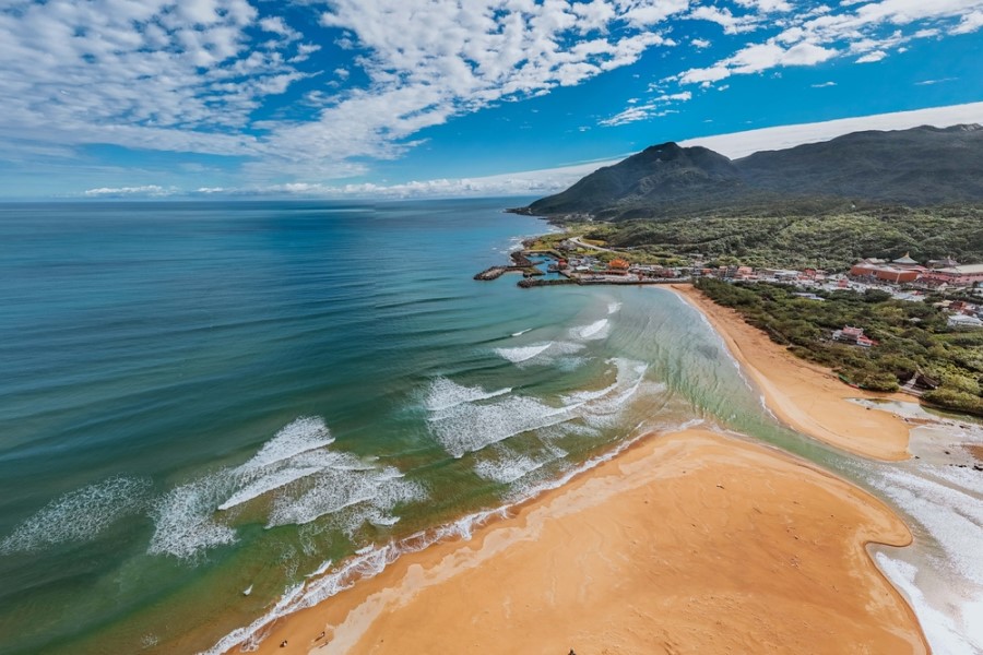 Ein goldgelber Sandstrand liegt an einer breiten Bucht mit sanften Wellen, im Hintergrund erstrecken sich grüne Hügel unter einem blauen Himmel mit weißen Wolken.