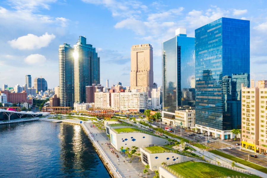 Eine moderne Stadtansicht mit hohen Glasgebäuden liegt an einem Flussufer mit Promenade, kleineren Häusern und grünen Bereichen im Vordergrund.