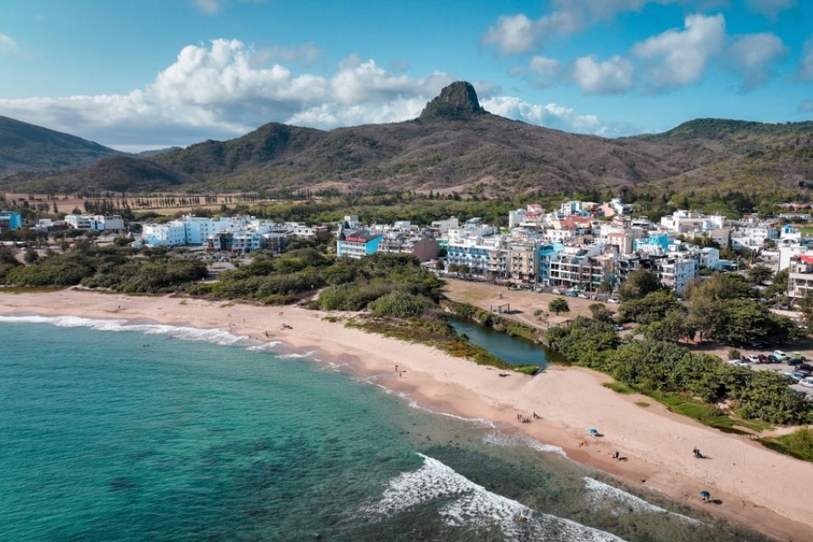 Eine kleine Stadt mit bunten Gebäuden liegt an einem Sandstrand mit türkisblauem Wasser, dahinter erheben sich grüne Hügel und ein markanter Felsgipfel.
