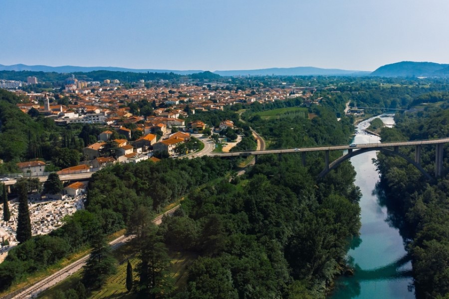 Eine Stadt mit orangefarbenen Dächern liegt inmitten grüner Hügel, eine Brücke führt über einen Fluss, der sich durch die Landschaft schlängelt.