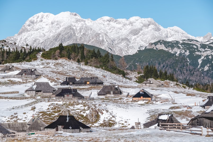 Mehrere Holzhütten stehen auf einer leicht verschneiten Bergwiese, im Hintergrund ragen schneebedeckte Gipfel unter blauem Himmel auf.