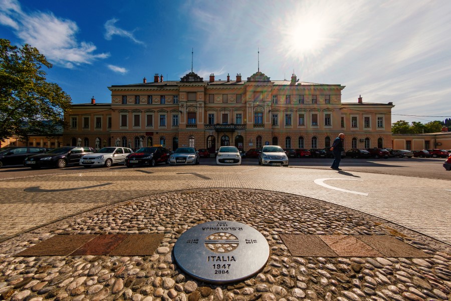 Ein großes historisches Gebäude mit geparkten Autos davor steht auf einem Platz mit einem runden Steinmosaik im Vordergrund, das eine Metallplakette mit Gravuren zeigt.