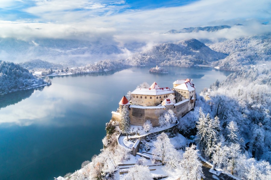 Eine Burg mit roten Dächern steht auf einem verschneiten Hügel über einem See, der von schneebedeckten Wäldern und Bergen umgeben ist.
