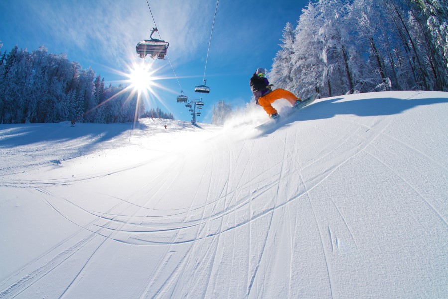 Eine Person fährt mit einem Snowboard eine verschneite Piste hinab, während im Hintergrund Skilifte und die tief stehende Sonne zu sehen sind.