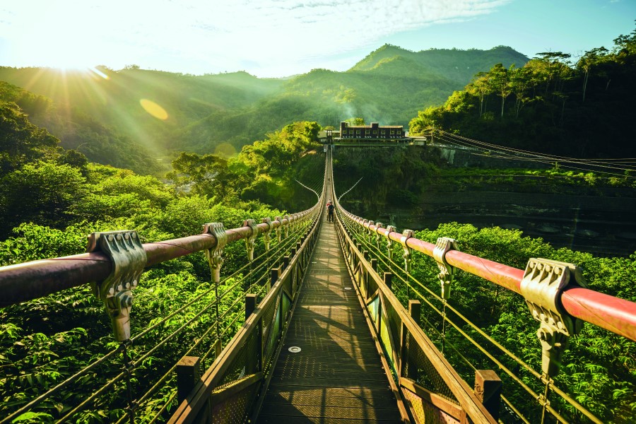 Eine lange Hängebrücke führt über dicht bewachsene grüne Hügel zu einem Gebäude, im Hintergrund scheint die Sonne über die Berge.