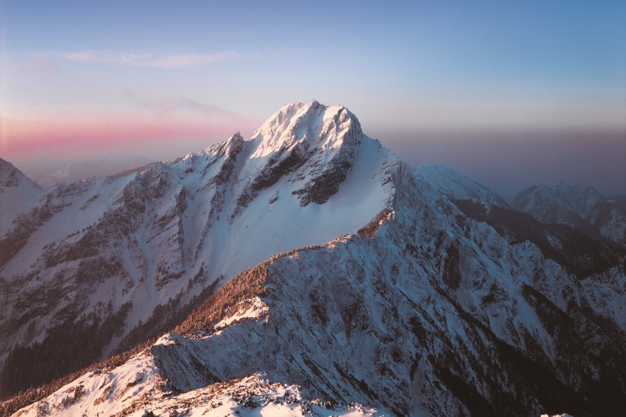 Ein schneebedeckter Berggipfel ragt über eine Reihe weiterer verschneiter Hänge, im Hintergrund färbt sich der Himmel leicht rosa und blau.