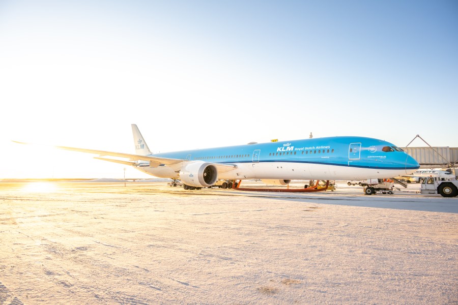 Ein blau-weißes Flugzeug steht auf einem sonnenbeschienenen Rollfeld, im Hintergrund sind Bodenfahrzeuge und ein Terminalgebäude zu sehen.