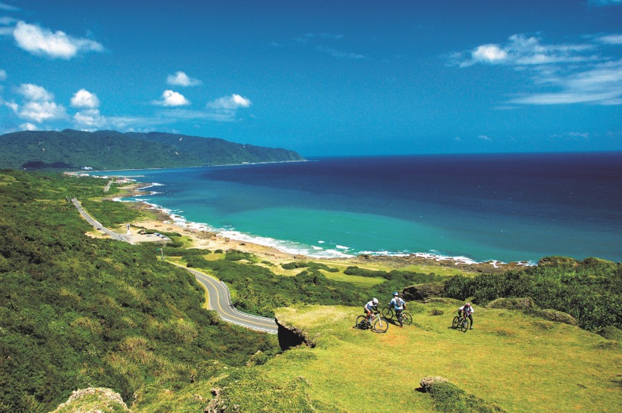 Drei Radfahrer fahren über eine grüne Küstenlandschaft mit gewundener Straße, im Hintergrund liegen das blaue Meer und bewaldete Hügel.