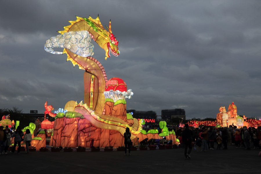 Ein großer leuchtender Drachen aus bunten Laternen steht auf einer beleuchteten Plattform, umgeben von Menschen und weiteren Lichtfiguren unter einem bewölkten Himmel.