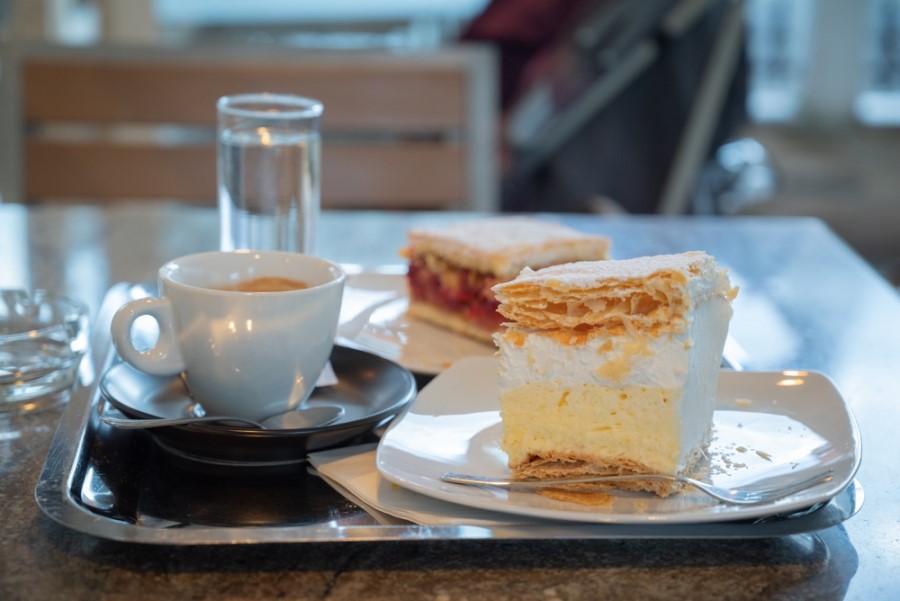 Eine Tasse Kaffee steht neben einem Glas Wasser und zwei Stück Kuchen auf einem Tablett, eines mit heller Creme und Blätterteig, das andere mit roter Füllung.