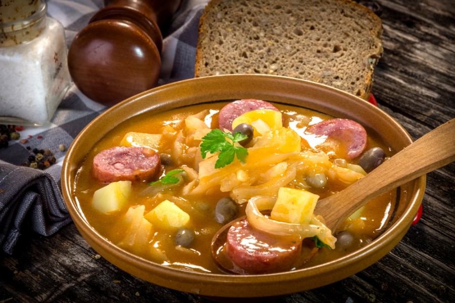 Ein Teller mit Eintopf aus Wurstscheiben, Kartoffelstücken, Kohl und Gewürzen steht auf einem Holztisch, daneben liegen Brotscheiben und Gewürzstreuer.