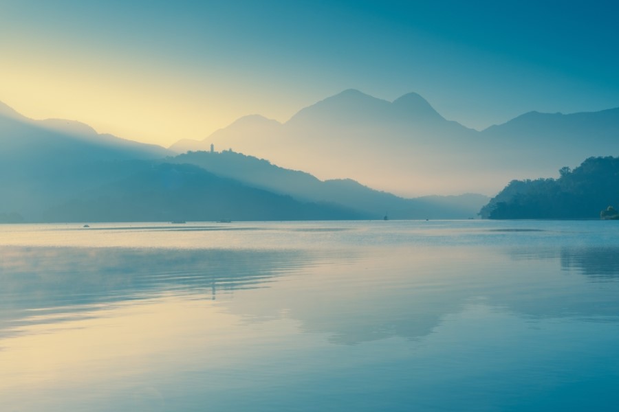 Ein ruhiger See spiegelt sanfte Hügel und ferne Berge im weichen Licht eines nebligen Morgens.