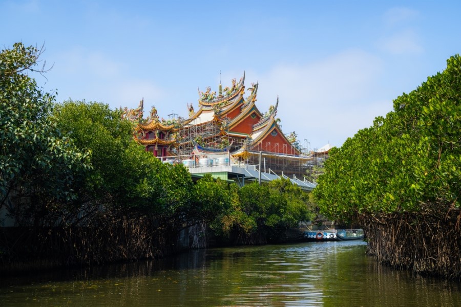 Ein bunt verziertes Tempelgebäude mit geschwungenen Dächern steht hinter dichten grünen Bäumen an einem ruhigen Wasserkanal.