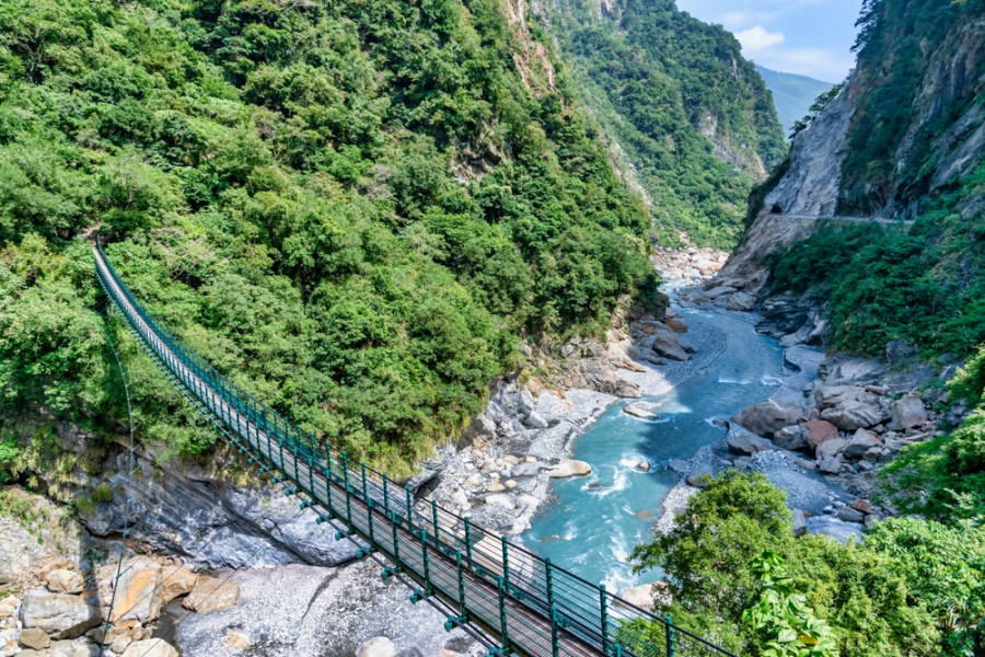 Eine Hängebrücke überspannt eine tiefe Schlucht mit türkisfarbenem Fluss, umgeben von steilen Felsen und dicht bewachsenen grünen Hängen.