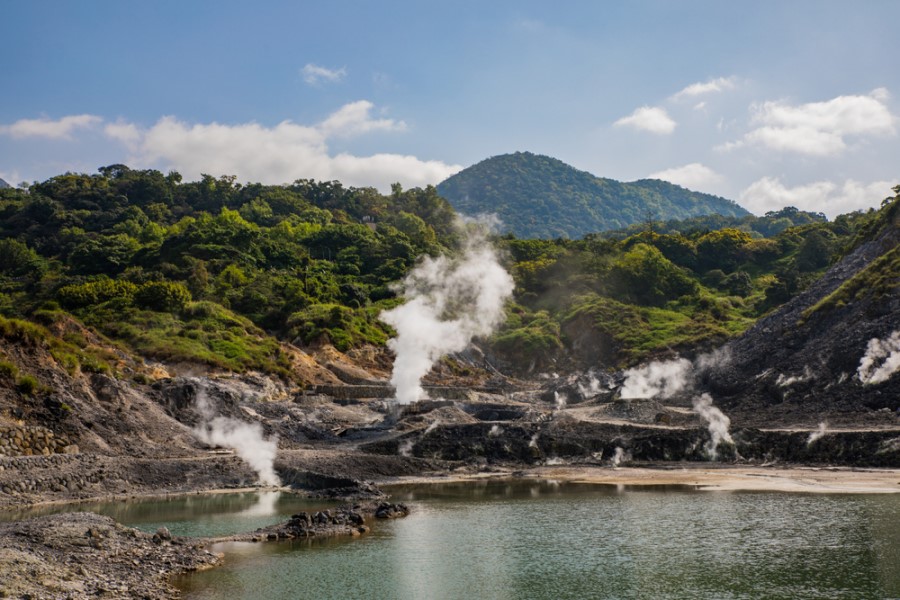 Ein geothermisches Gebiet mit dampfenden Quellen liegt vor grünen Hügeln, der Dampf steigt aus mehreren Stellen rund um ein Wasserbecken auf.