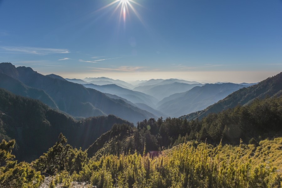 Eine Berglandschaft mit grünen Hängen und bewaldeten Tälern erstreckt sich unter einem klaren Himmel, in dem die Sonne hell über den Gipfeln steht.